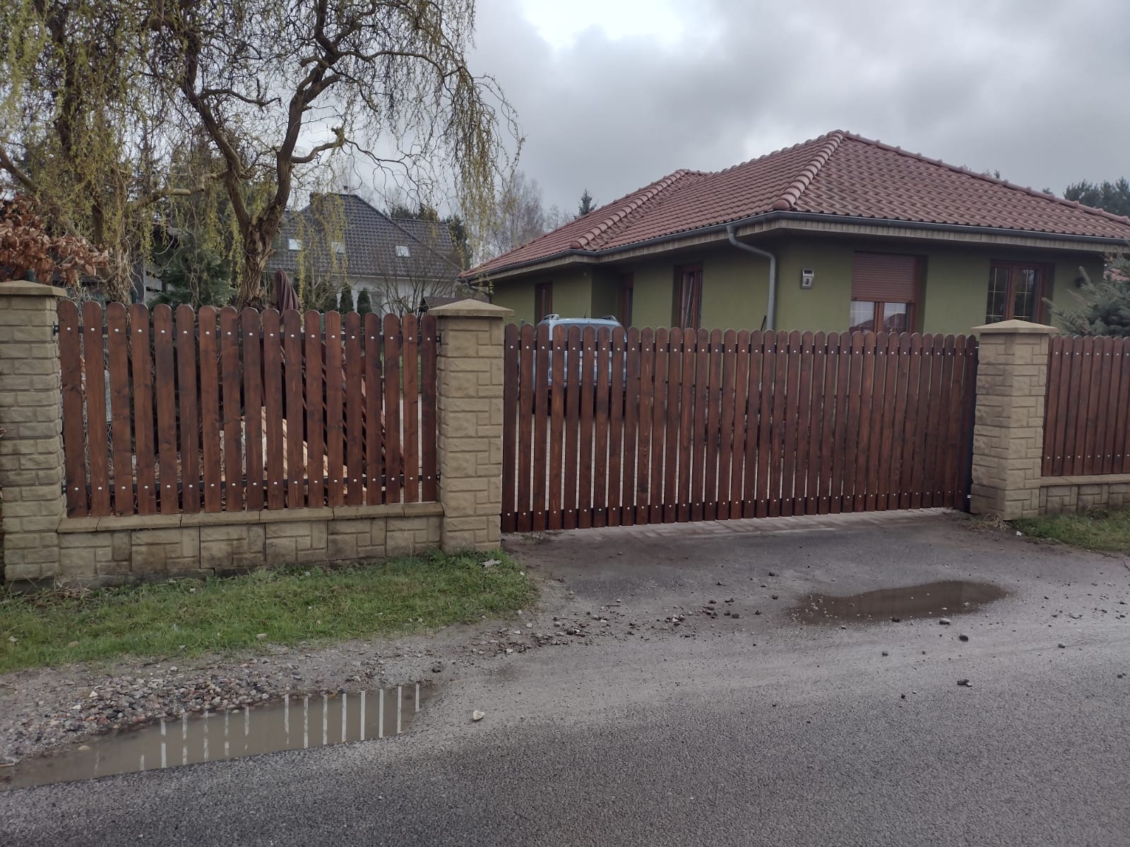 Drewniana brama wjazdowa z pionowych desek, osadzona na murowanych słupkach, przed domem z czerwoną dachówką, widok na kałużę na asfalcie.