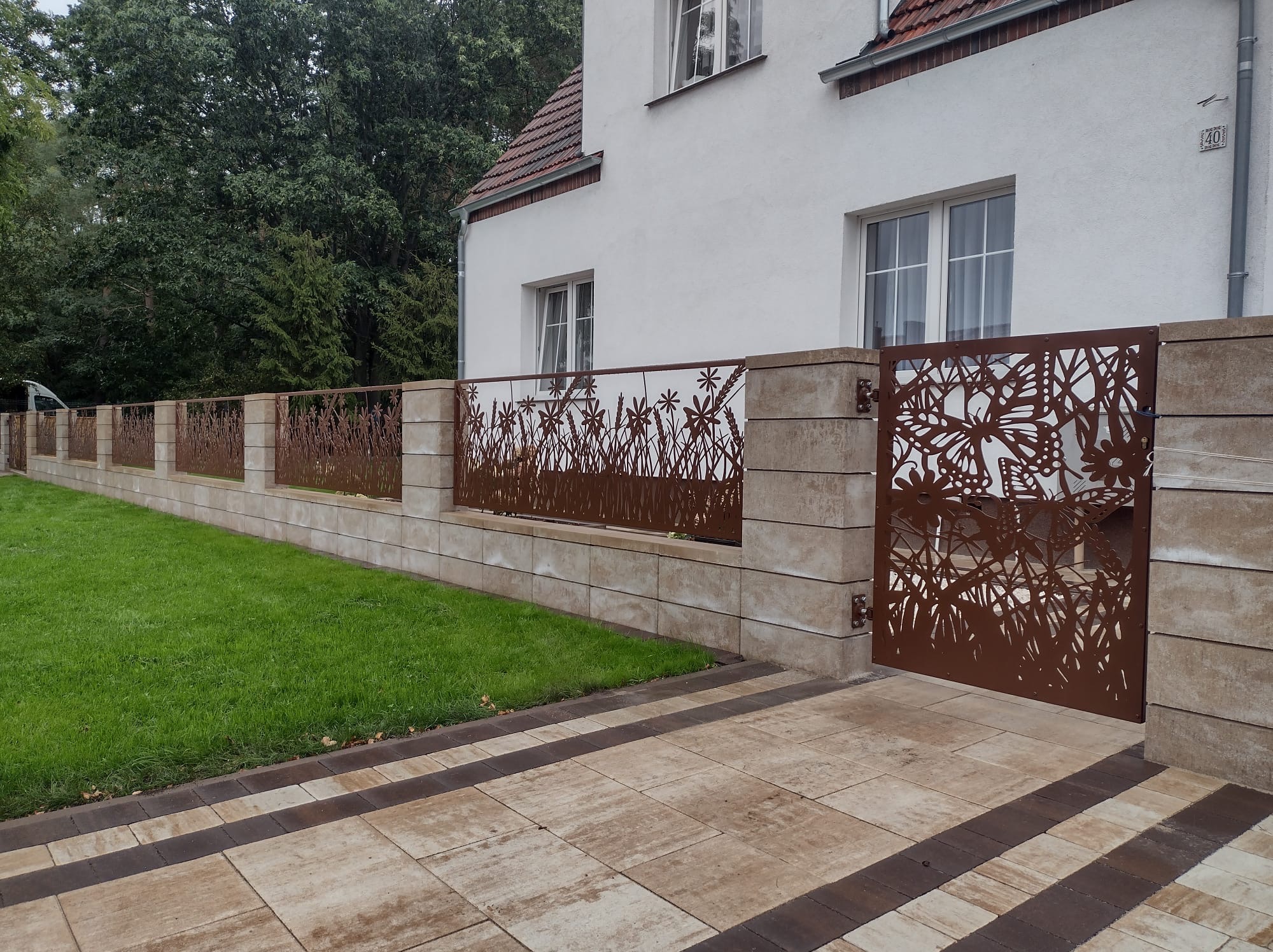 Nowoczesne ogrodzenie z metalową bramą wjazdową z motywem roślinnym i motyli, osadzone na niskim murku z bloków betonowych, na tle domu z ogrodem.