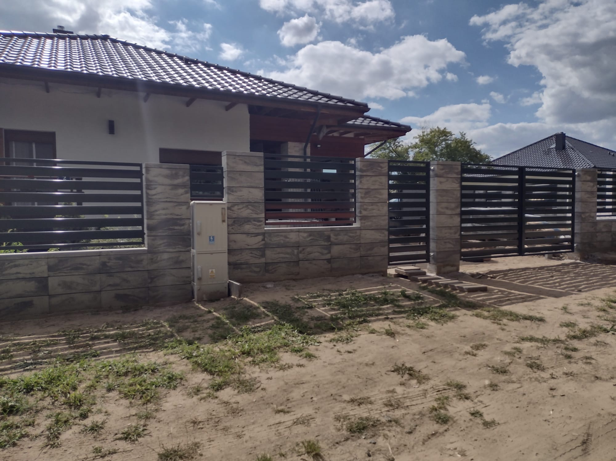 Nowoczesne ogrodzenie panelowe z elementami imitującymi kamień, skrzynka elektryczna na słupku, dom jednorodzinny w tle, widok z poziomu gruntu.