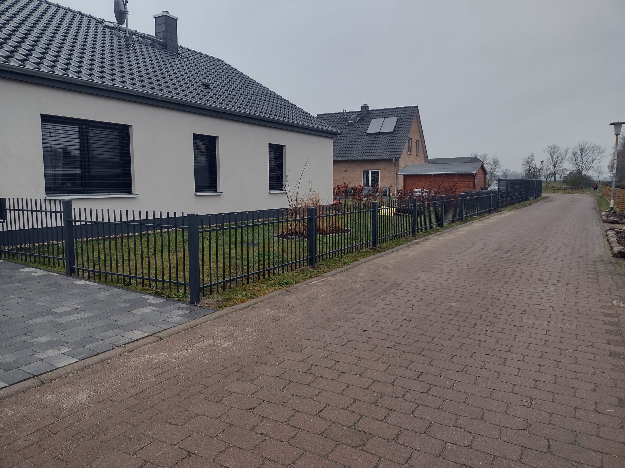 Szary metalowy płot sztachetowy oddziela posesję z białym domem od chodnika z czerwonej kostki brukowej w pochmurny dzień.