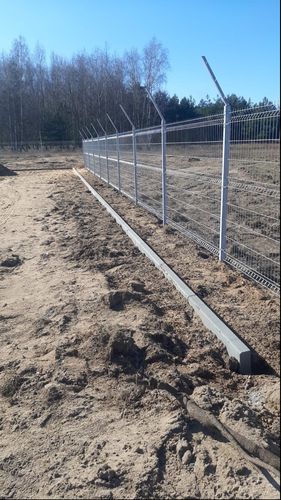 Długi odcinek ogrodzenia panelowego z drutem kolczastym na słupkach, zamontowany na betonowej podmurówce, z widocznym piaszczystym podłożem i lasem w tle.