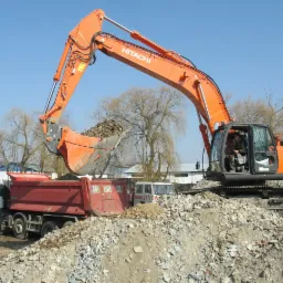 Pomarańczowa koparka Hitachi załadowuje gruz do czerwonej ciężarówki na tle błękitnego nieba. Widoczny operator w kabinie koparki.