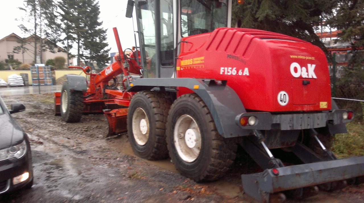 Czerwony równiarka O&K F156.6A w Lublinie, widok z tyłu. Maszyna stoi na błotnistym terenie, obok zaparkowany samochód. Widoczne drzewa i budynki w tle.