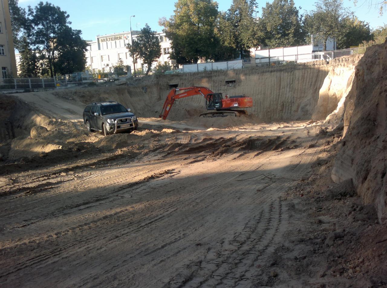 Pomarańczowa koparka Hitachi 390 w wykopie ziemnym, obok szary samochód terenowy. W tle budynek z napisem MULTIDRUK.