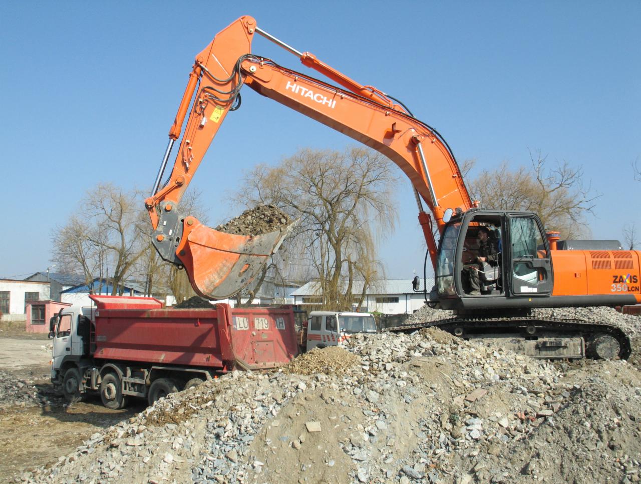 Pomarańczowa koparka Hitachi załadowuje gruz do czerwonej ciężarówki na tle błękitnego nieba. Widoczny operator w kabinie koparki.
