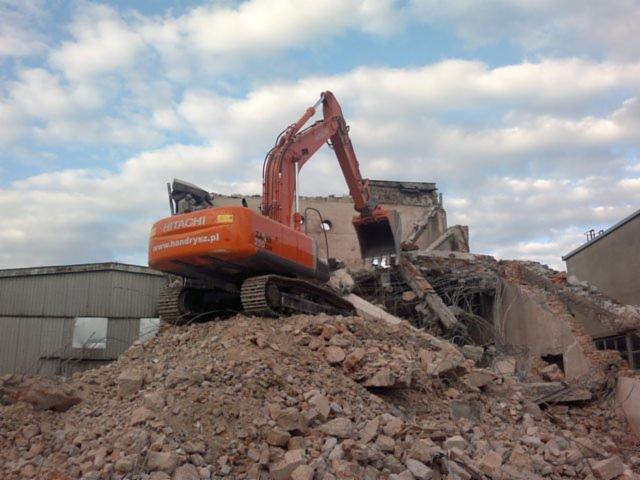 Pomarańczowa koparka Hitachi na gruzowisku, w tle fragment zburzonego budynku i pochmurne niebo. Widoczny adres strony internetowej na maszynie.