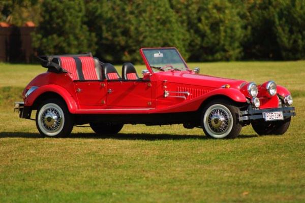 Czerwony, zabytkowy kabriolet z białymi oponami, zaparkowany na zielonym trawniku w słoneczny dzień. Widok z boku, z detalami chromowanych elementów i tablicy rejestracyjnej.