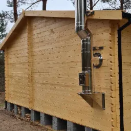 Zewnętrzny, metalowy przewód dymowy zamontowany na bocznej ścianie drewnianego domku letniskowego, widoczne elementy mocujące i wyczystka.