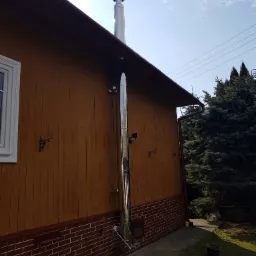 Zewnętrzny, stalowy komin zamontowany przy drewnianym domu z ceglaną podmurówką, widoczny fragment dachu i nieba.