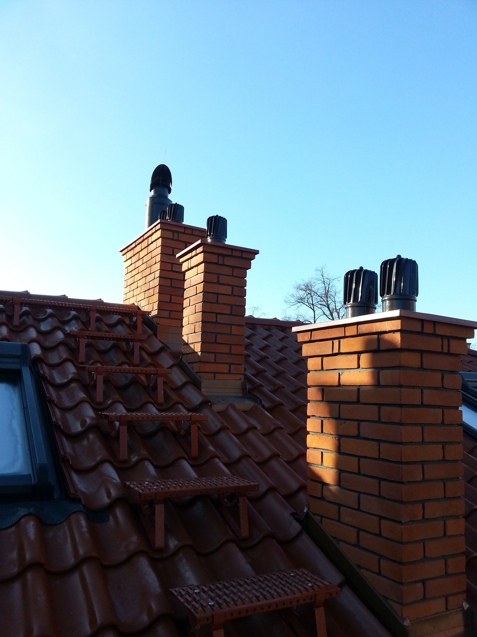 Czerwona dachówka z zamontowanymi stopniami kominiarskimi prowadzącymi do trzech kominów z cegły klinkierowej zakończonych czarnymi nasadami kominowymi, błękitne niebo w tle.