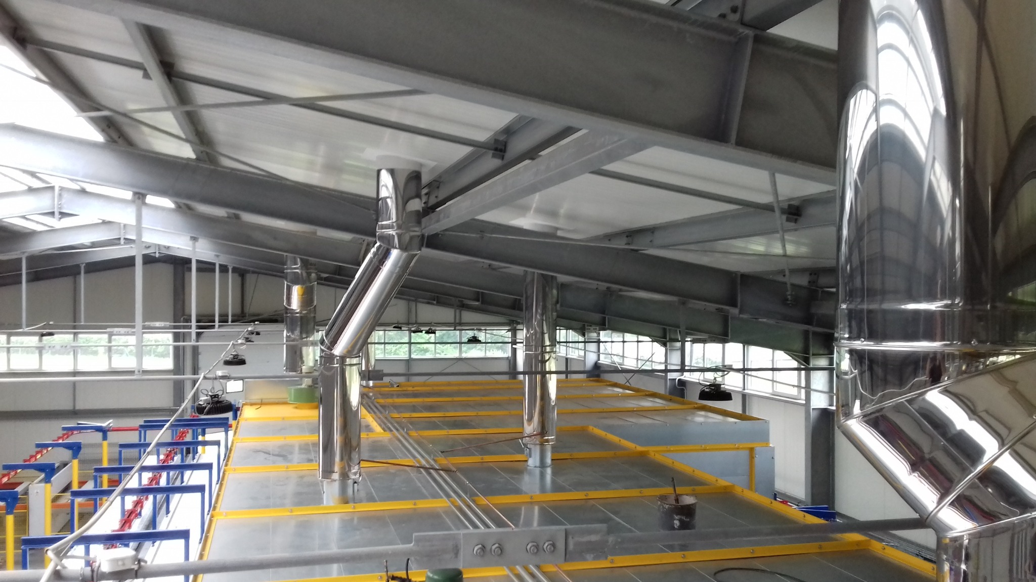 Wnętrze hali przemysłowej z systemem wentylacji, widoczne metalowe rury i kratownice pod sufitem, żółte obramowania na podłodze i elementy linii produkcyjnej w tle.