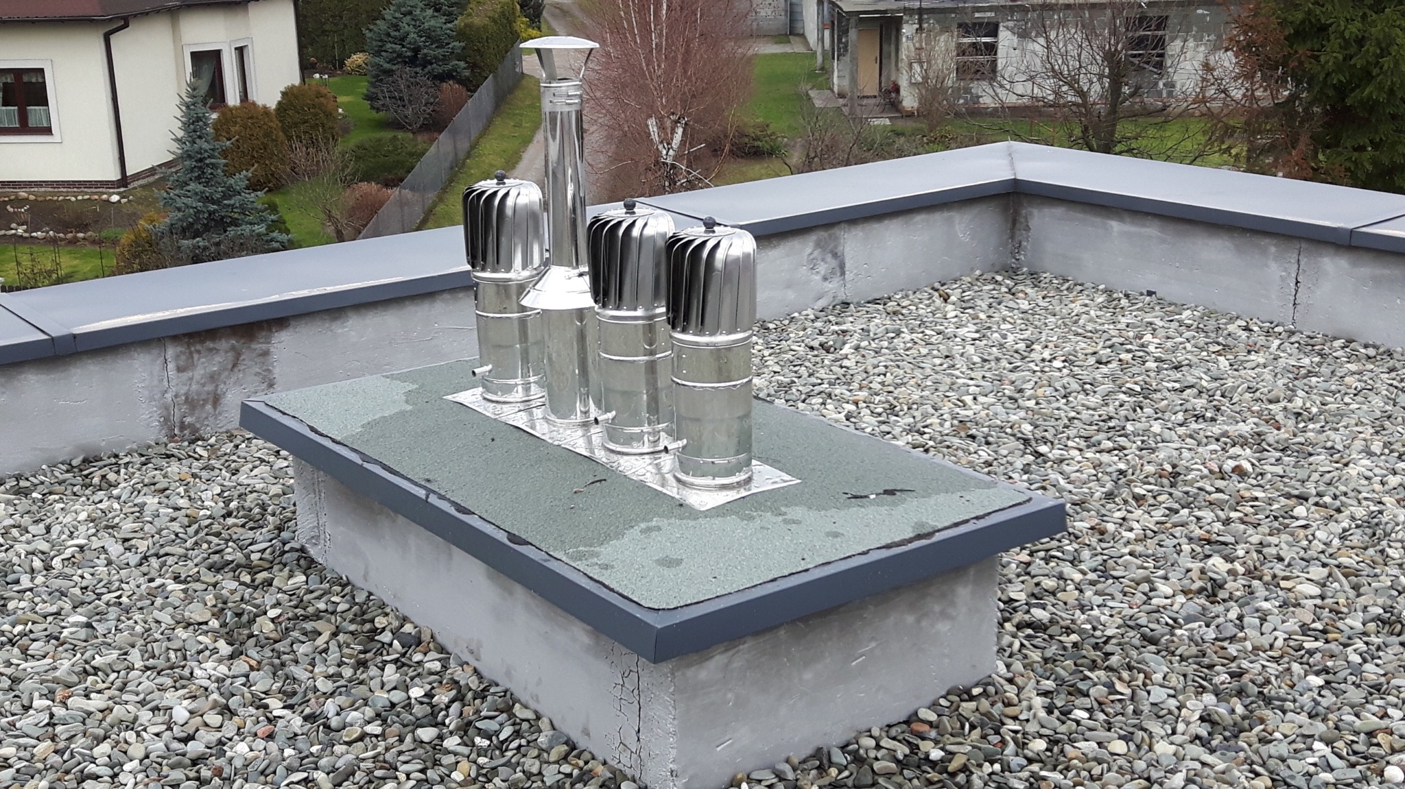 Cztery metalowe zakończenia przewodów wentylacyjnych na płaskim dachu pokrytym kamykami, z widocznymi budynkami w tle.