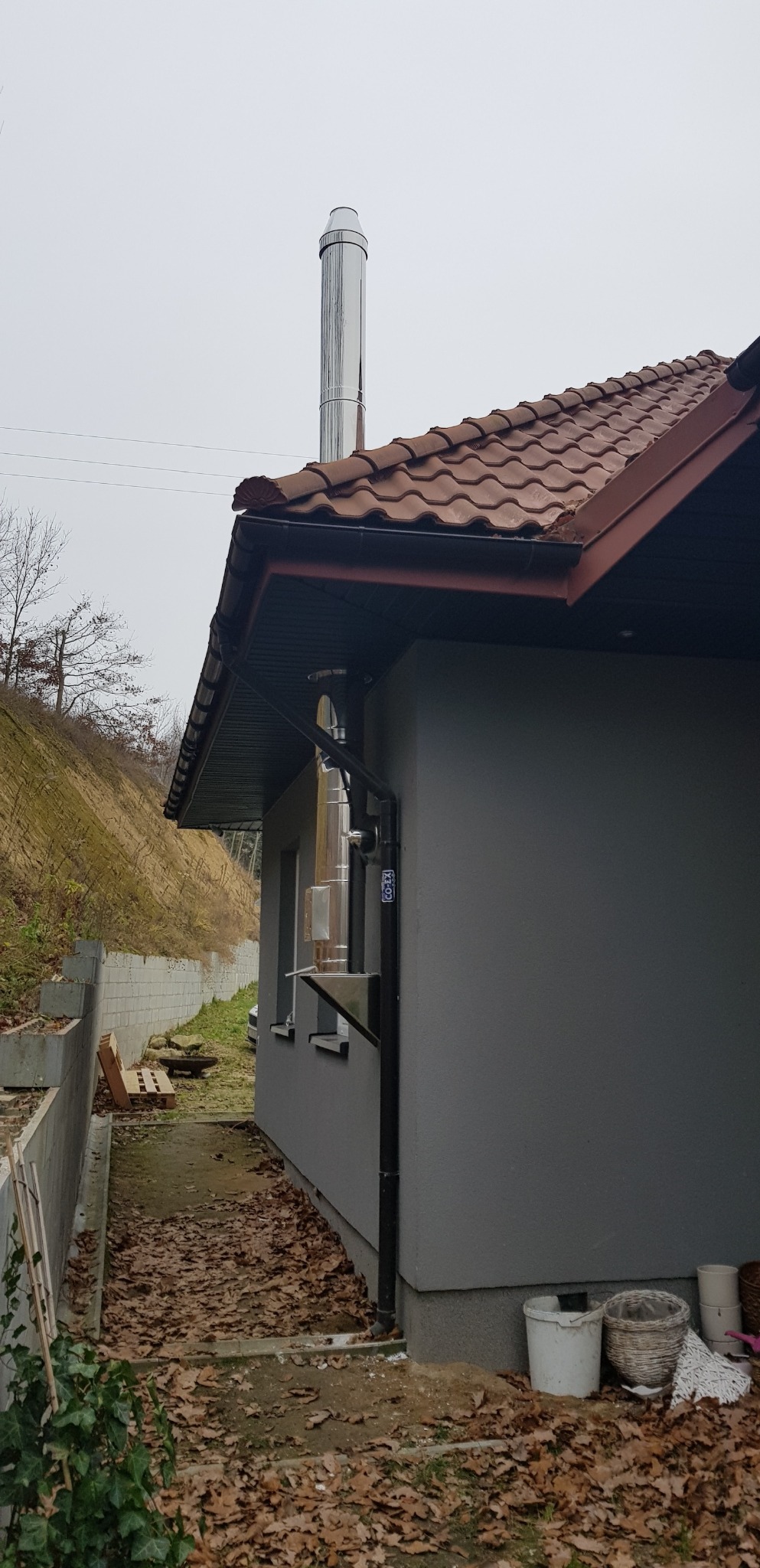 Zewnętrzny, stalowy system odprowadzania spalin, zamocowany do ściany budynku z brązowym dachem, widoczny w pochmurny dzień.