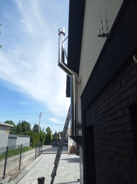 Zewnętrzny, stalowy przewód kominowy zamontowany przy ścianie budynku z drabiny opierającej się o ścianę, widok z dołu na błękitne niebo.