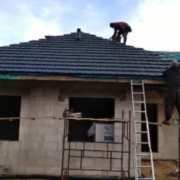 Montaż czarnej dachówki na nowym domu w trakcie budowy, widoczni dekarze pracujący na dachu i rusztowaniach, część dachu pokryta zieloną membraną.