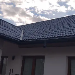 Nowoczesny, ciemny dach z blachy trapezowej, z zamontowanymi rynnami, widok z dołu na tle pochmurnego nieba, fragment rusztowania z lewej strony.