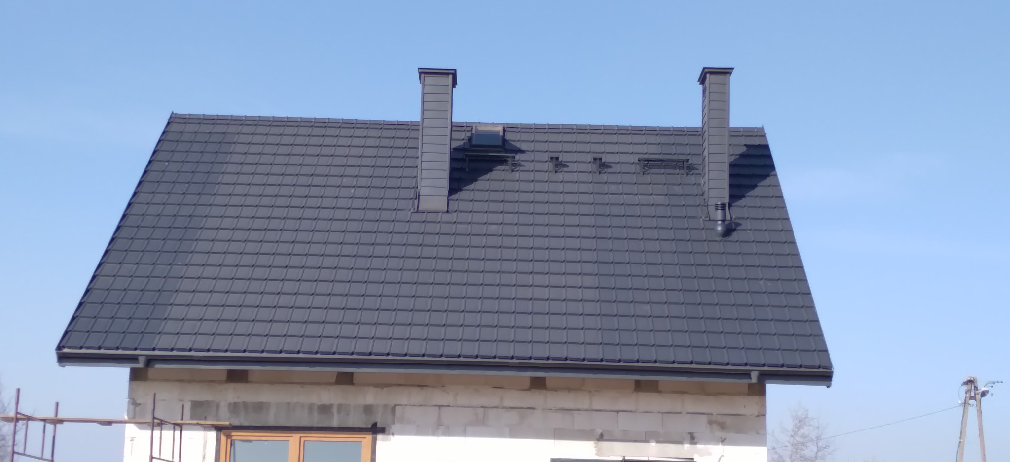 Nowy, grafitowy dach z dwoma kominami i oknem dachowym na surowym budynku w trakcie budowy, na tle błękitnego nieba.