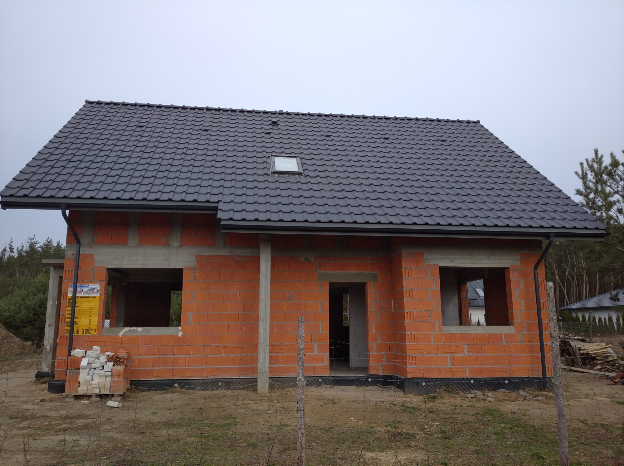 Dom w trakcie budowy z częściowo ukończonym dachem z ciemnej dachówki i zamontowanymi rynnami, widoczne niezamurowane otwory okienne i drzwiowe.