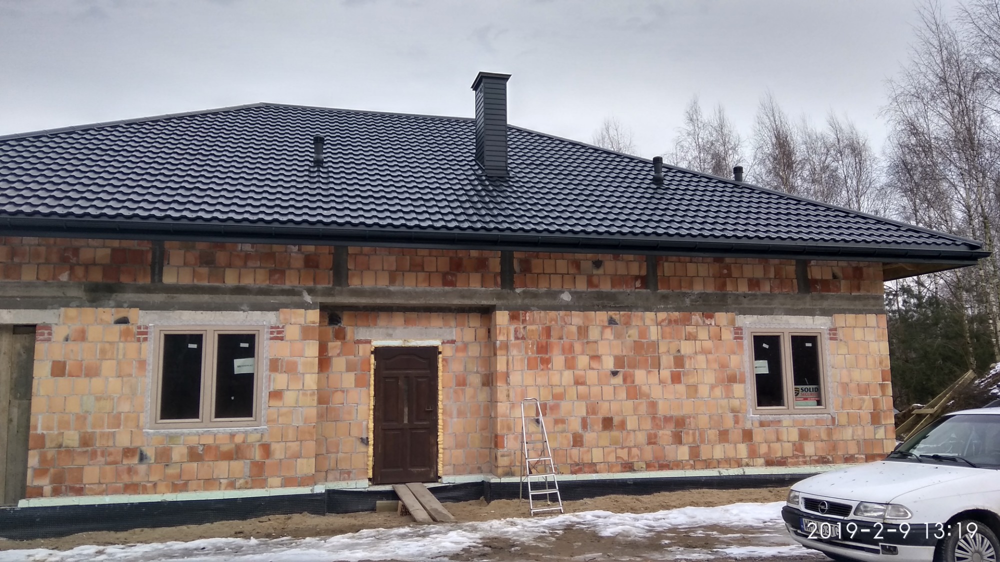 Niedokończony budynek z ciemną blachodachówką, widoczne rynny, komin i fragment białego samochodu na pierwszym planie. Na ścianach surowy mur z cegły, okna wstawione, ale nieotynkowane. Przed...