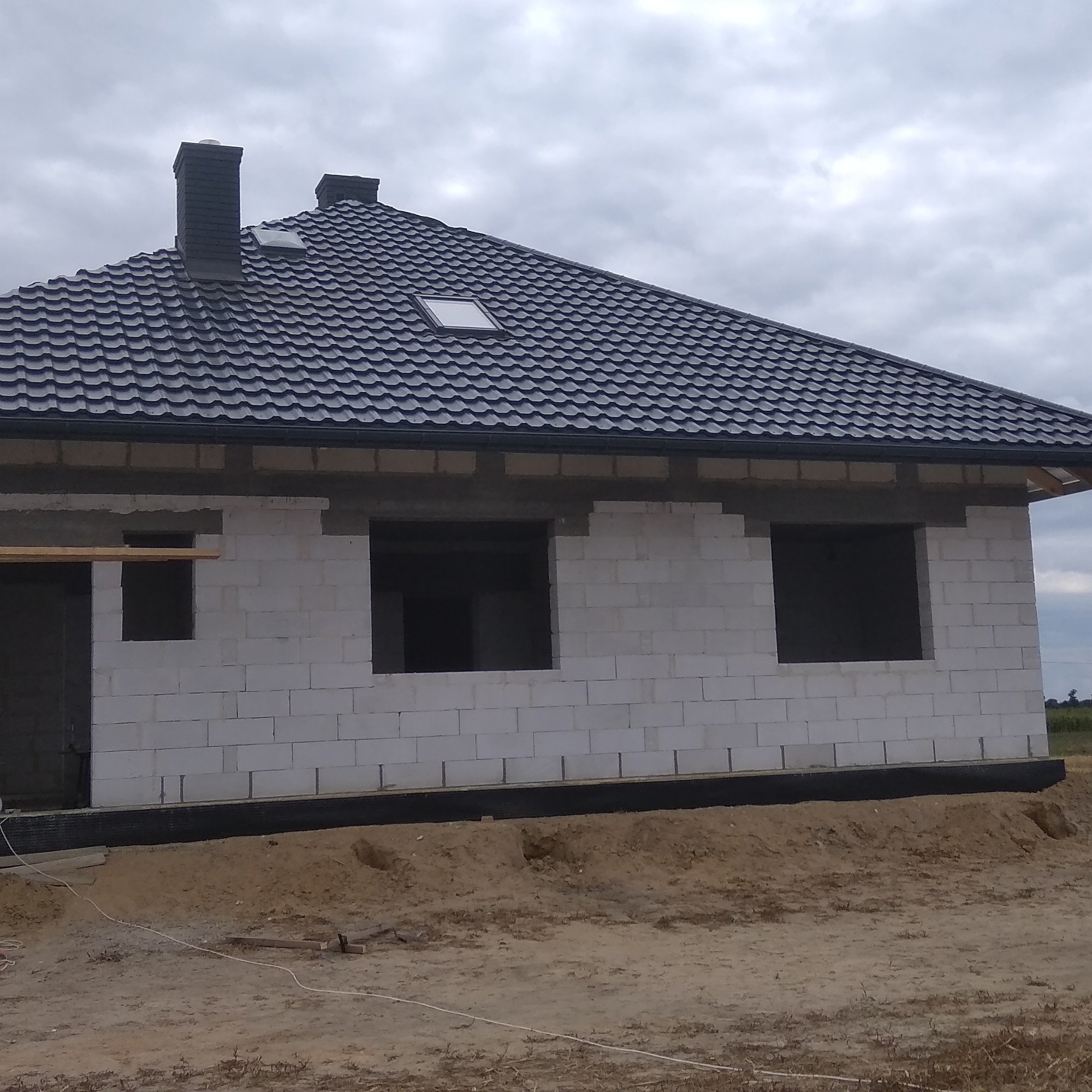 Surowy stan domu z czarnym dachem z dachówki modułowej, widoczne okno dachowe i komin, ściany z betonu komórkowego bez okien i drzwi, teren wokół domu w trakcie przygotowania.