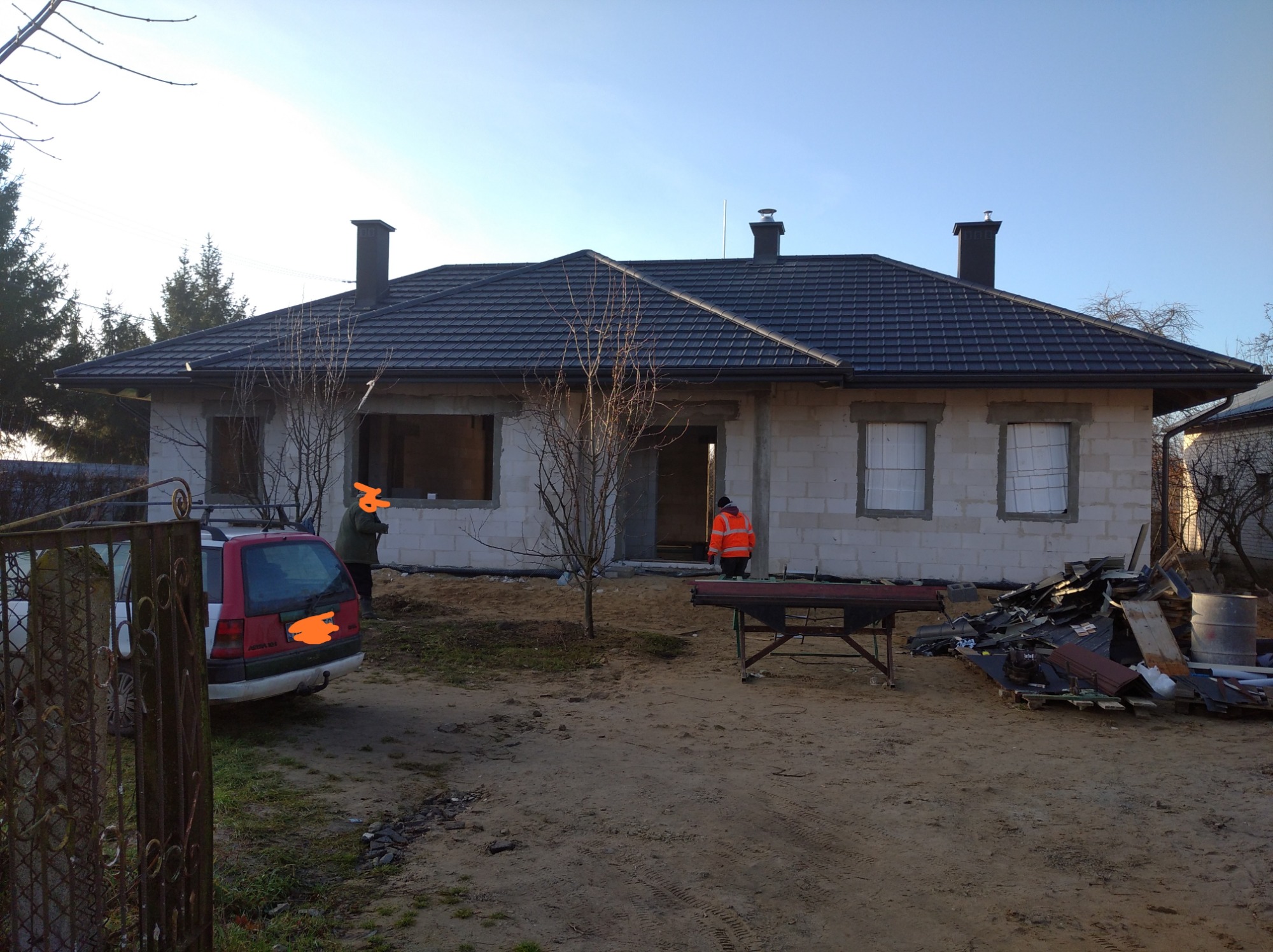 Dom w trakcie budowy z ciemną dachówką, widoczny samochód, narzędzia i materiały budowlane na posesji.