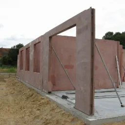 Wznoszenie ścian domu modułowego z betonu architektonicznego na fundamencie, widoczne podpory stabilizujące, w tle budynek gospodarczy z czerwonej cegły i żuraw budowlany.