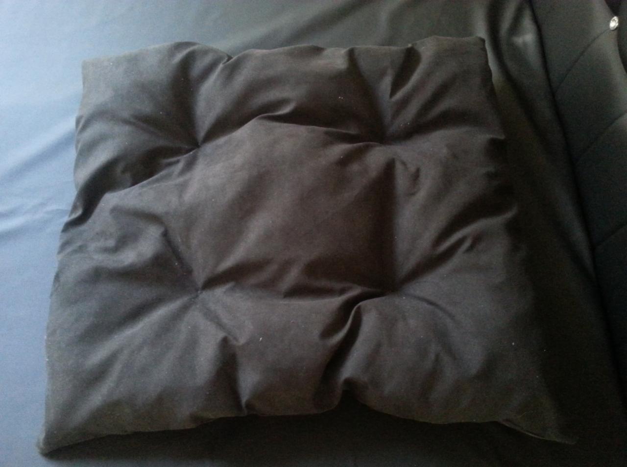 Ciemna, kwadratowa poduszka dekoracyjna z pikowaniem, leżąca na niebieskiej tkaninie. Widoczna faktura materiału i delikatne załamania światła.