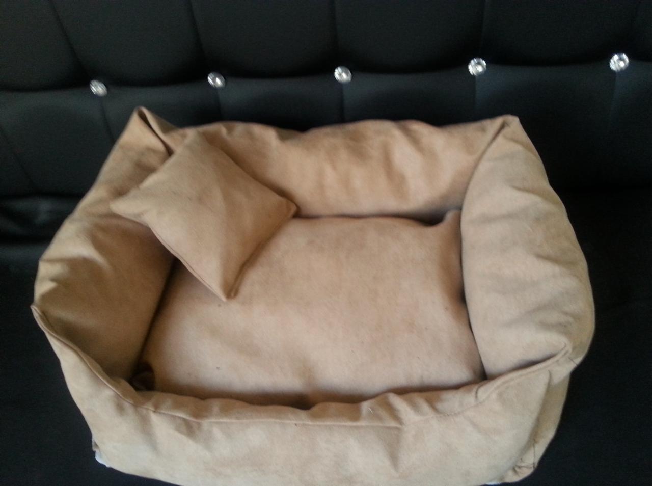 Beżowe legowisko dla zwierząt z poduszką, uszyte w Żaganiu. Miękkie, tapicerowane posłanie na tle czarnej sofy.