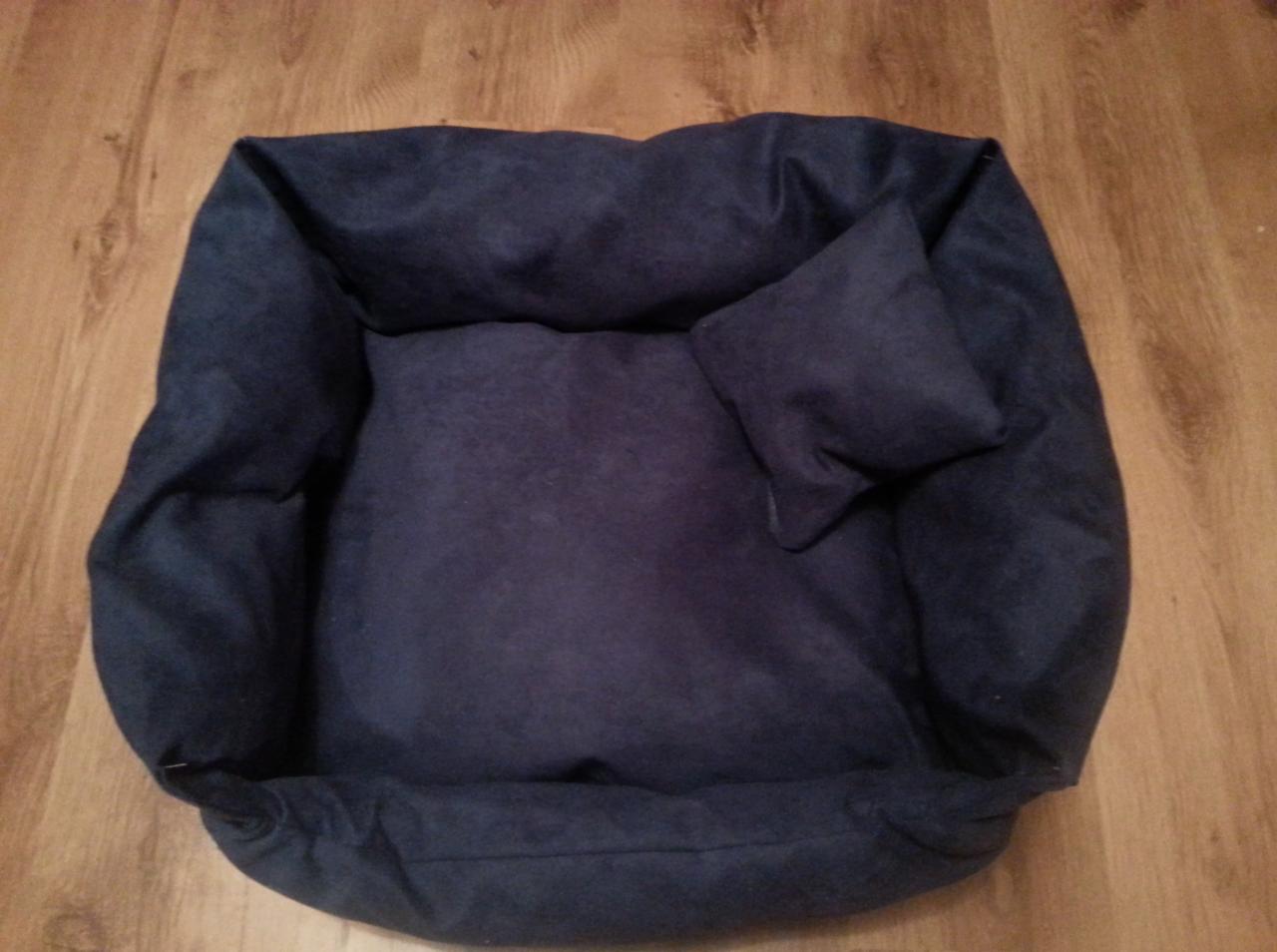 Granatowe, pluszowe legowisko dla psa z małą poduszką, leżące na drewnianej podłodze. Widok z góry, miękkie oświetlenie.