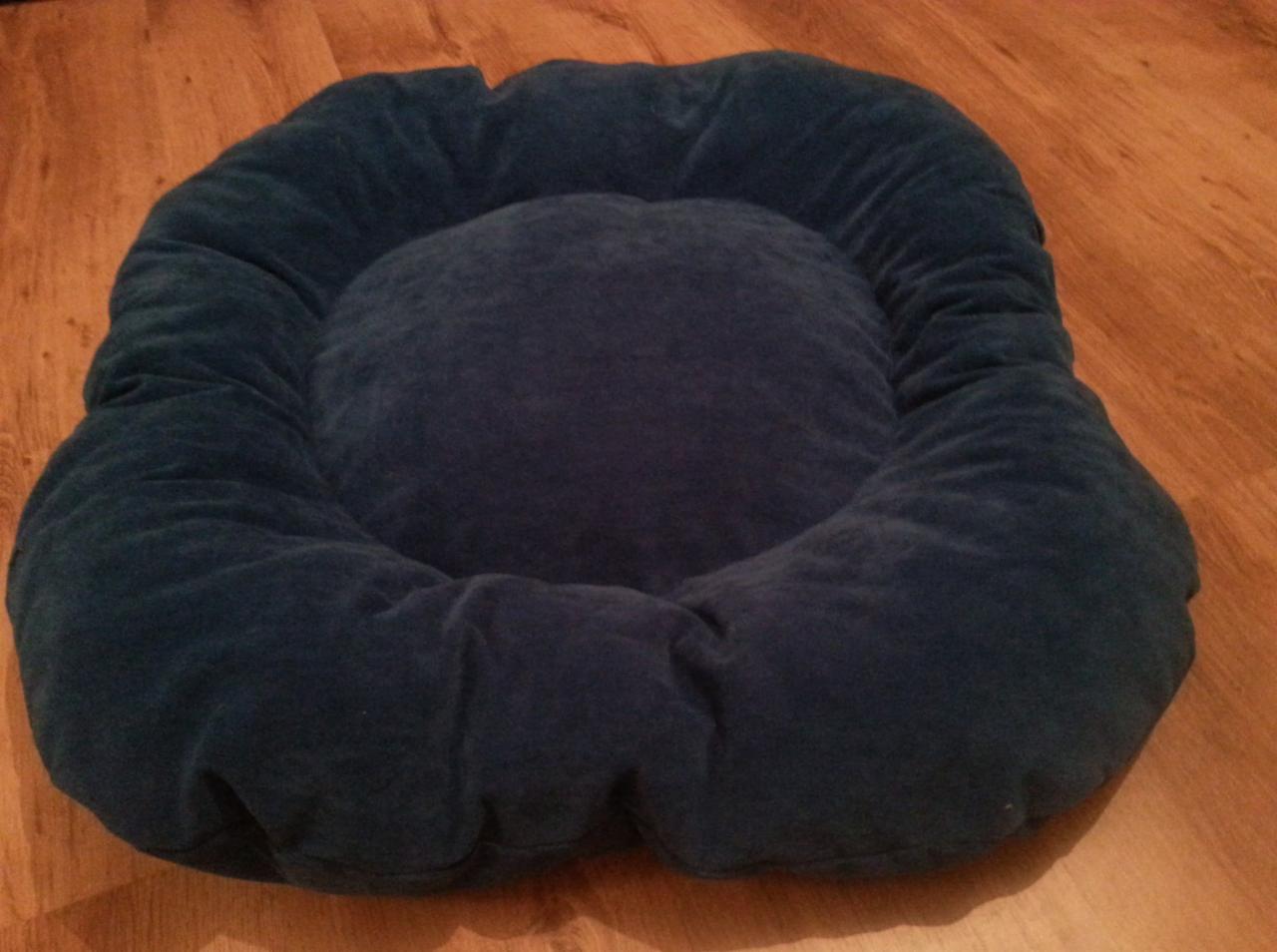Granatowe, welurowe legowisko dla zwierząt na drewnianej podłodze. Miękkie, okrągłe boki i centralna poduszka. Widok z góry, naturalne światło.