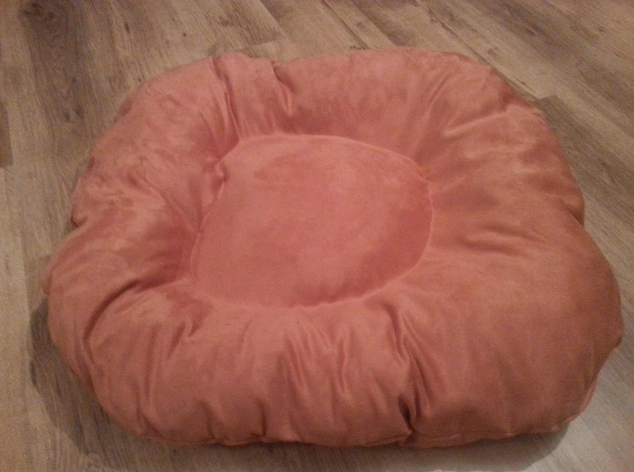 Puszysta, kwadratowa poduszka w odcieniach brzoskwini leży na drewnianej podłodze. Widoczne delikatne marszczenia materiału i centralne wgłębienie.