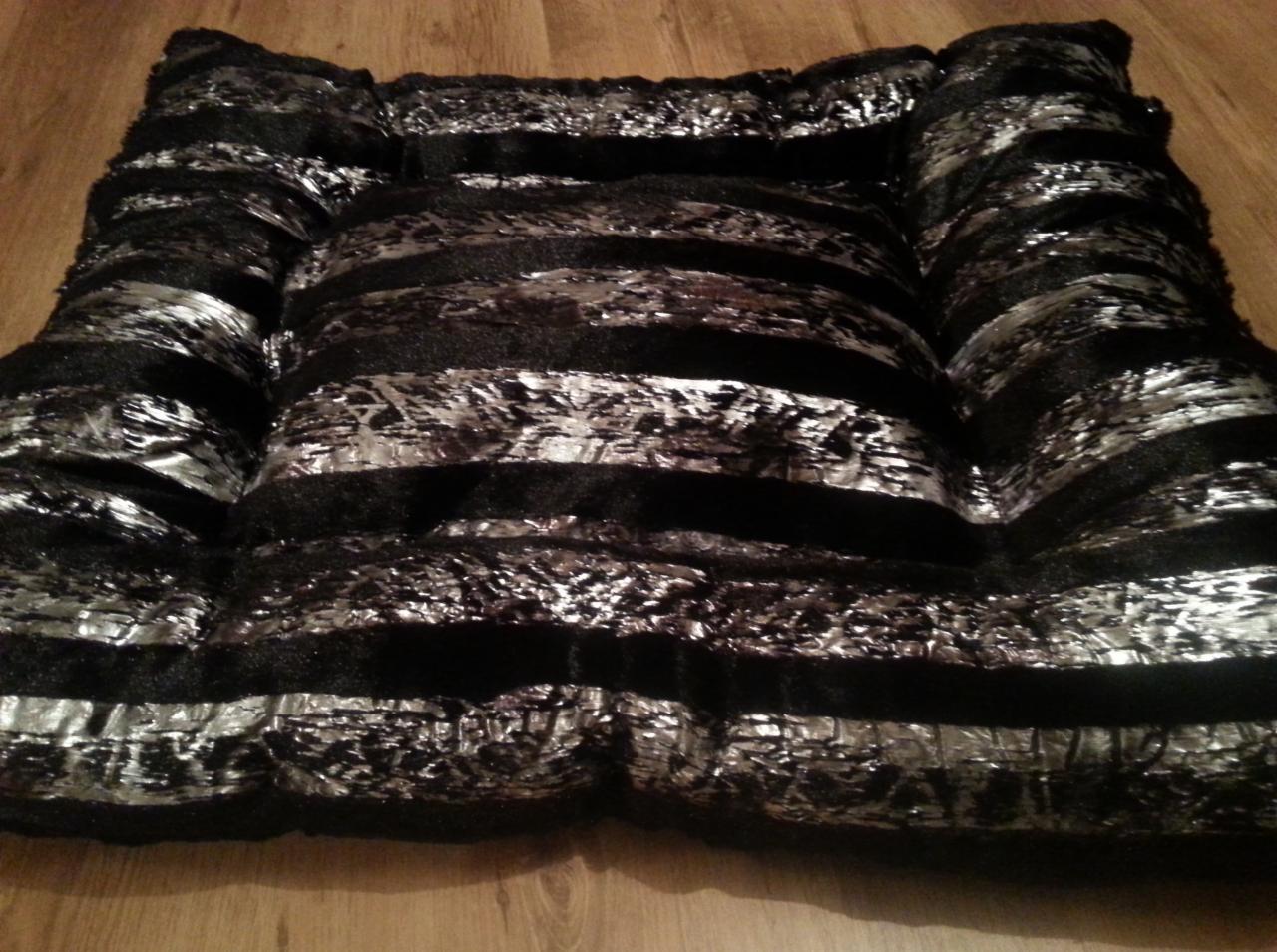 Czarno-srebrna, błyszcząca poduszka/materac w paski, leżąca na drewnianej podłodze. Widoczne szwy i faktura materiału. Nietypowe połączenie faktur i kolorów.