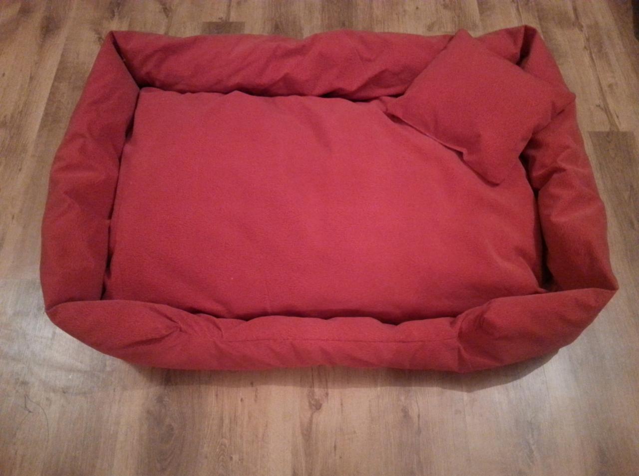 Czerwone legowisko dla psa z małą poduszką, wykonane z miękkiego materiału, leży na drewnianej podłodze. Widok z góry, proste szwy.