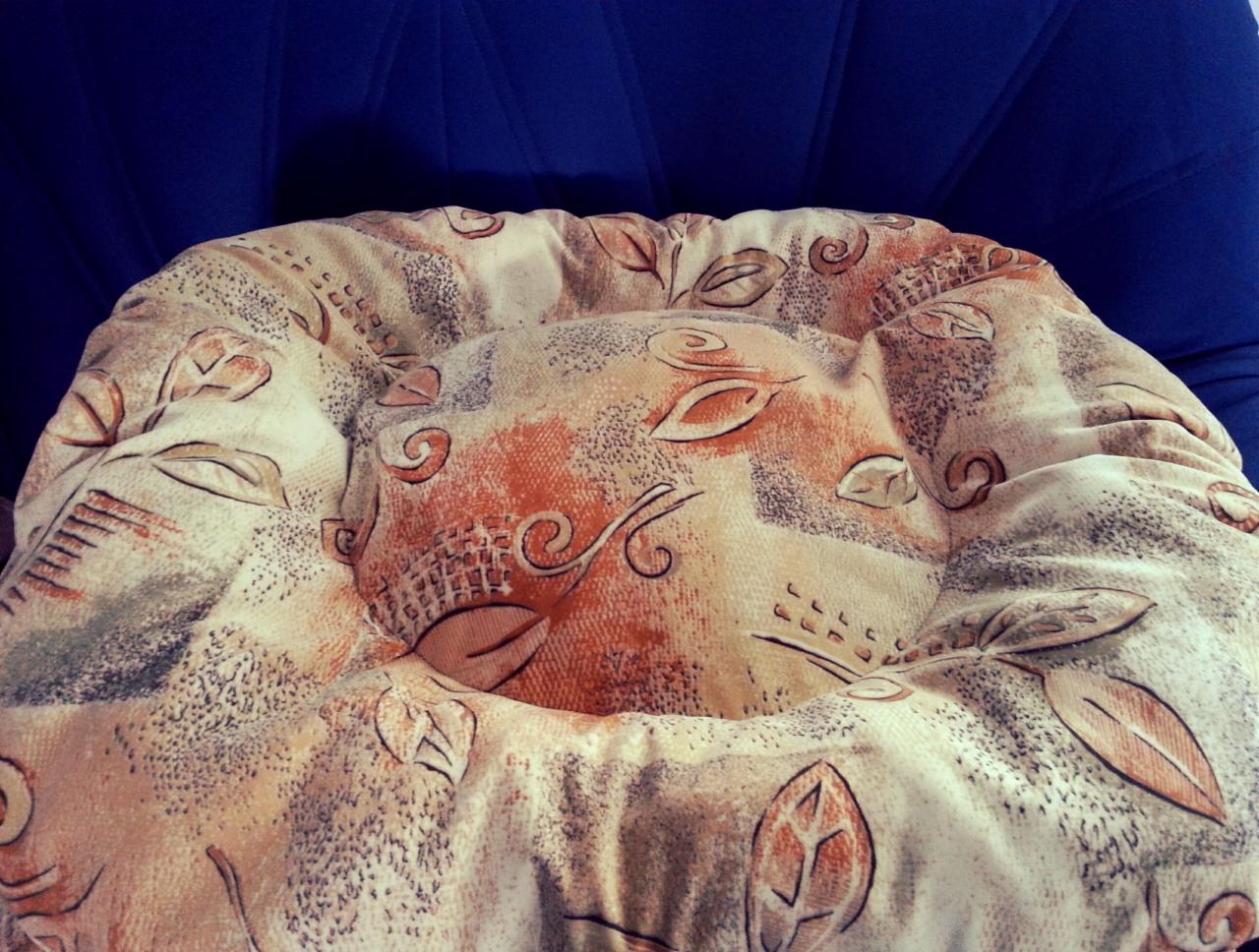 Okrągła poduszka na krzesło z wzorem liści i spiral na kremowym tle, leżąca na niebieskim krześle. Widok z góry, miękkie światło.