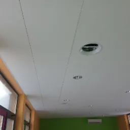 Biały sufit panelowy z wbudowanym oświetleniem punktowym i okrągłym nawiewnikiem, widok z dołu. Pomarańczowe ściany z oknami i klimatyzator w tle.