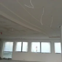 Wykończony sufit z profilami gipsowymi i elementami oświetlenia w nowym wnętrzu. Okna zabezpieczone folią malarską. Widoczne kable oświetleniowe.