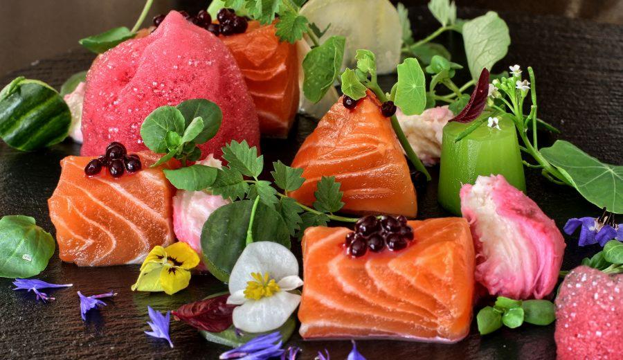 Elegancka kompozycja z kawałków łososia, różowych i zielonych kostek, ozdobiona kwiatami i drobnymi owocami na ciemnym tle.