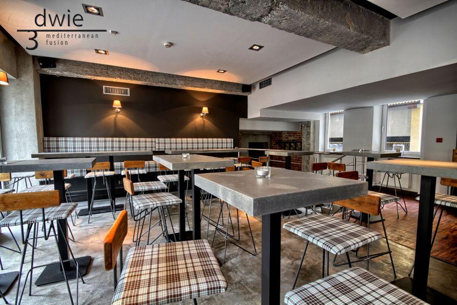 Wnętrze restauracji z wysokimi stolikami i krzesłami w kratę, betonowe blaty, surowy beton na suficie, czarna ściana z lampkami, logo 'dwie 3 mediterranean fusion' w rogu.