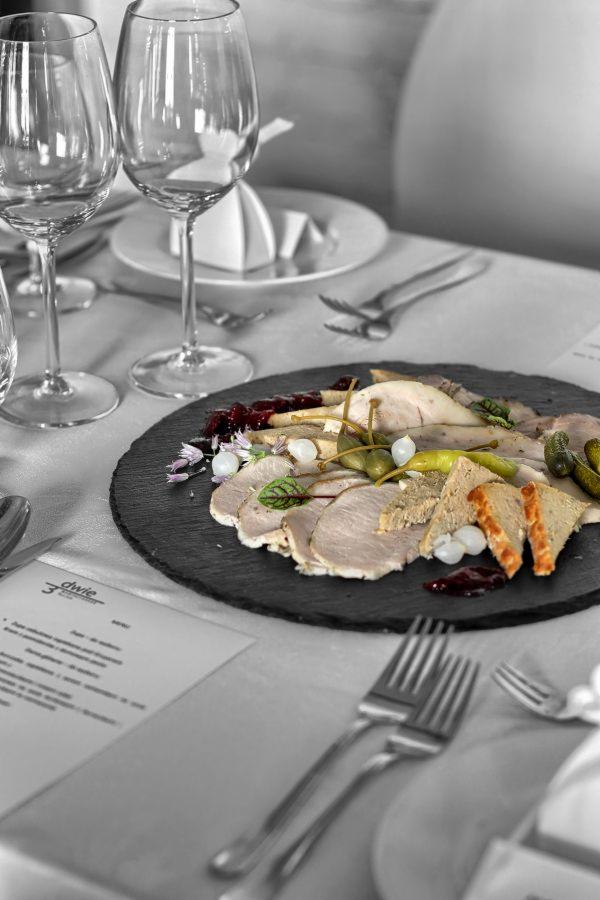 Elegancko podane plastry mięsa z kaparami, piklami, cebulkami i grzankami na czarnym, okrągłym talerzu, ustawionym na stole z nakryciem, obok stoją kieliszki i leży menu.