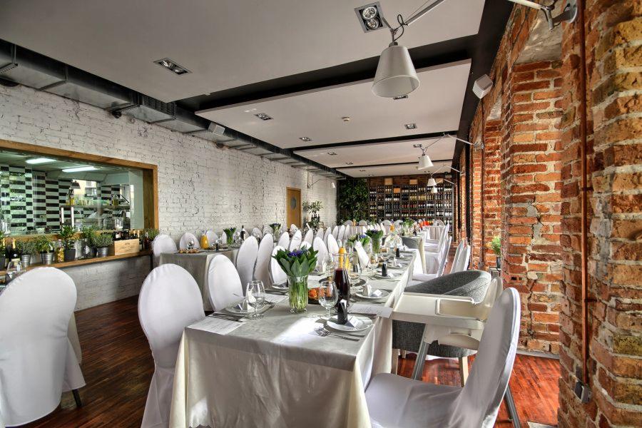 Ustawione stoły w sali bankietowej z ceglanymi ścianami i widokiem na kuchnię, przygotowane do organizacji imprezy firmowej.