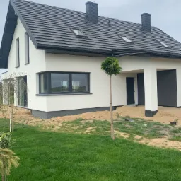 Biały dom jednorodzinny z szarą dachówką i czarnymi detalami wykończeniowymi, widoczny trawnik z młodymi drzewkami, teren wokół domu w trakcie prac wykończeniowych.