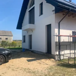 Nowoczesny dom z białą elewacją i czarnymi akcentami, częściowo osłonięty rusztowaniem, z widocznym samochodem i drugim budynkiem w tle.