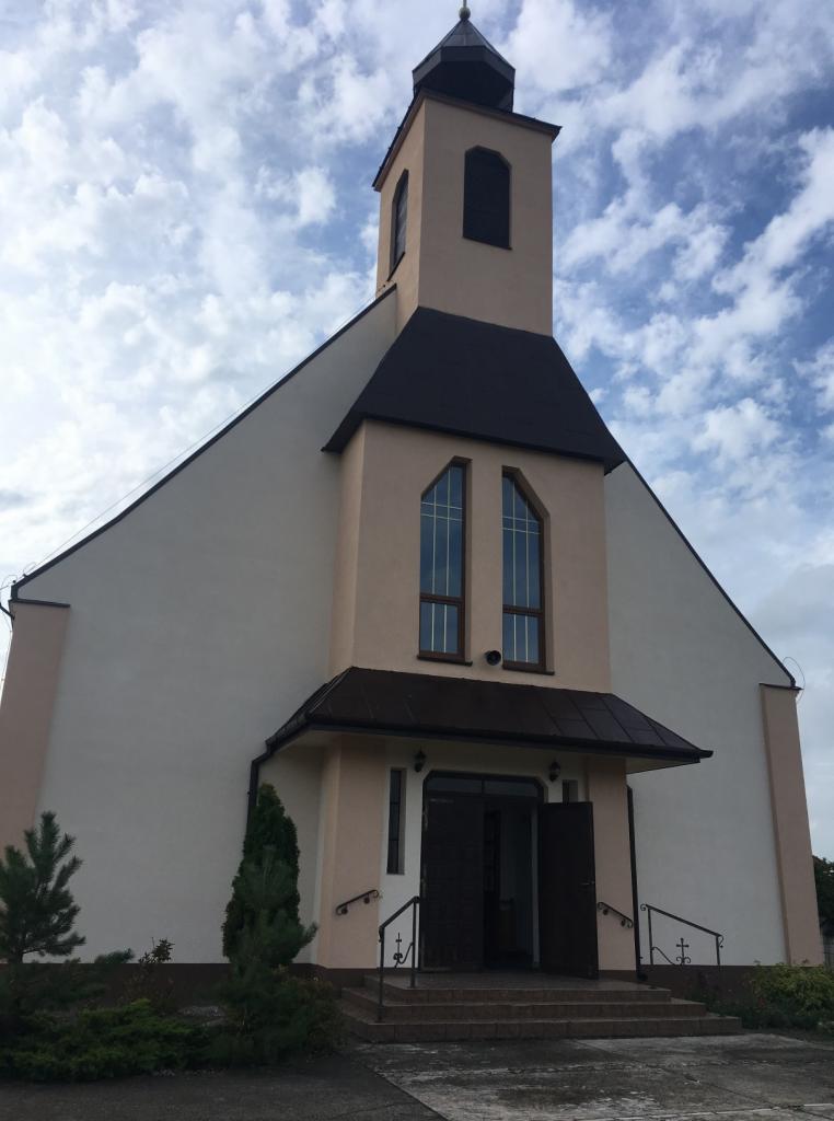 Odnowiona fasada kościoła w odcieniach beżu i bieli, z ciemnobrązowym dachem i wieżą, widok z dołu na tle pochmurnego nieba.