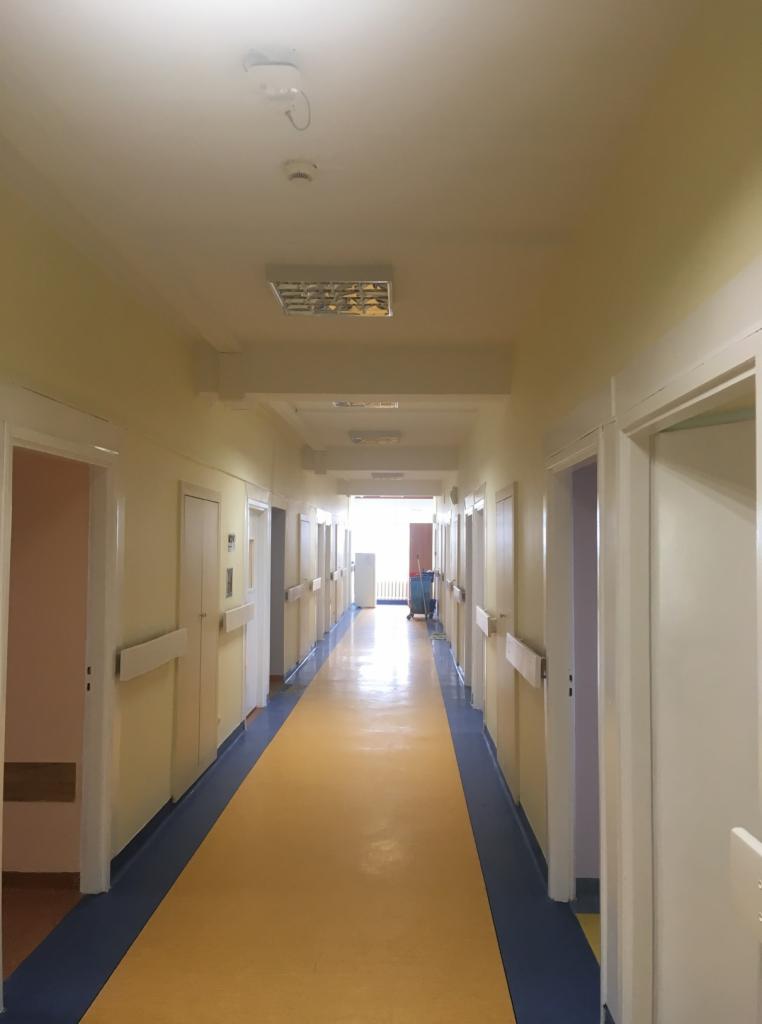 Długi, jasny korytarz w budynku użyteczności publicznej z żółtą podłogą i niebieskimi krawędziami, białe drzwi po obu stronach, widoczne oświetlenie sufitowe i wózek sprzątający na końcu.