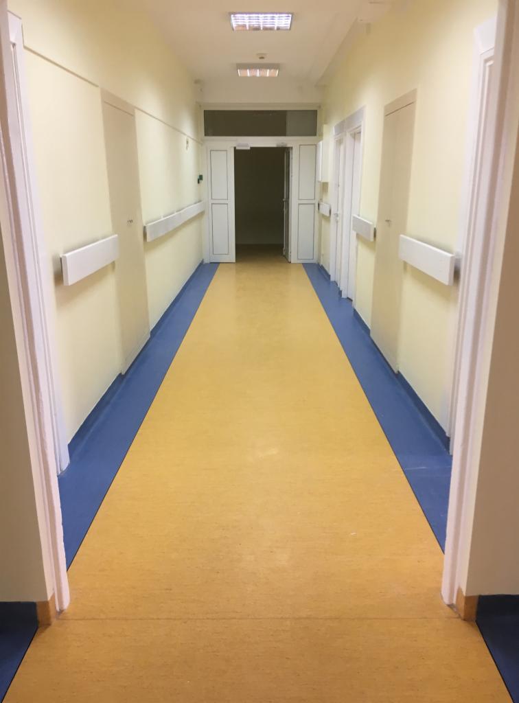 Długi, jasny korytarz w budynku użyteczności publicznej, z żółtą podłogą obramowaną niebieską listwą przypodłogową, białymi ścianami z zamontowanymi poręczami i szeregiem białych drzwi po obu...