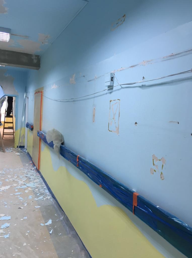 Korytarz w trakcie przygotowania do malowania w Gąsewie Poduchownym: ściany częściowo pokryte niebieską farbą, dolna część zabezpieczona żółtą folią, listwa przypodłogowa owinięta niebieską folią...