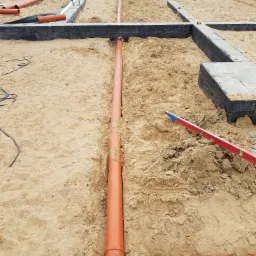 Fundamenty domu w trakcie budowy z widocznymi rurami kanalizacyjnymi w wykopie, poziomica leżąca na piasku i częściowo zalane betonem ściany fundamentowe.