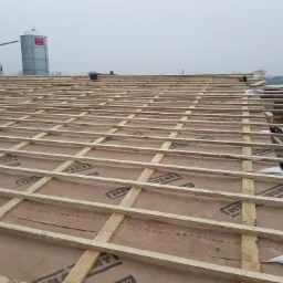 Konstrukcja drewniana dachu na brązowej membranie z widocznymi silosami w tle, prawdopodobnie na terenie gospodarstwa rolnego.