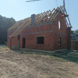 Wznoszenie domu z czerwonej cegły ceramicznej: widoczne ściany, otwory okienne i drzwiowe, komin oraz drewniana konstrukcja dachu w trakcie budowy, na tle błękitnego nieba i lasu, z żurawiem...