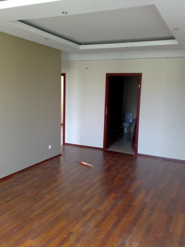 Pokój po remoncie z drewnianą podłogą, świeżo pomalowanymi ścianami w odcieniach beżu i wykończonym sufitem podwieszanym. Widoczna tuba silikonu na podłodze.